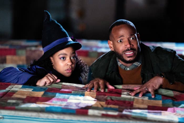 Marlon looking scared and hiding behind a table with a child who&#x27;s wearing a witch&#x27;s hat
