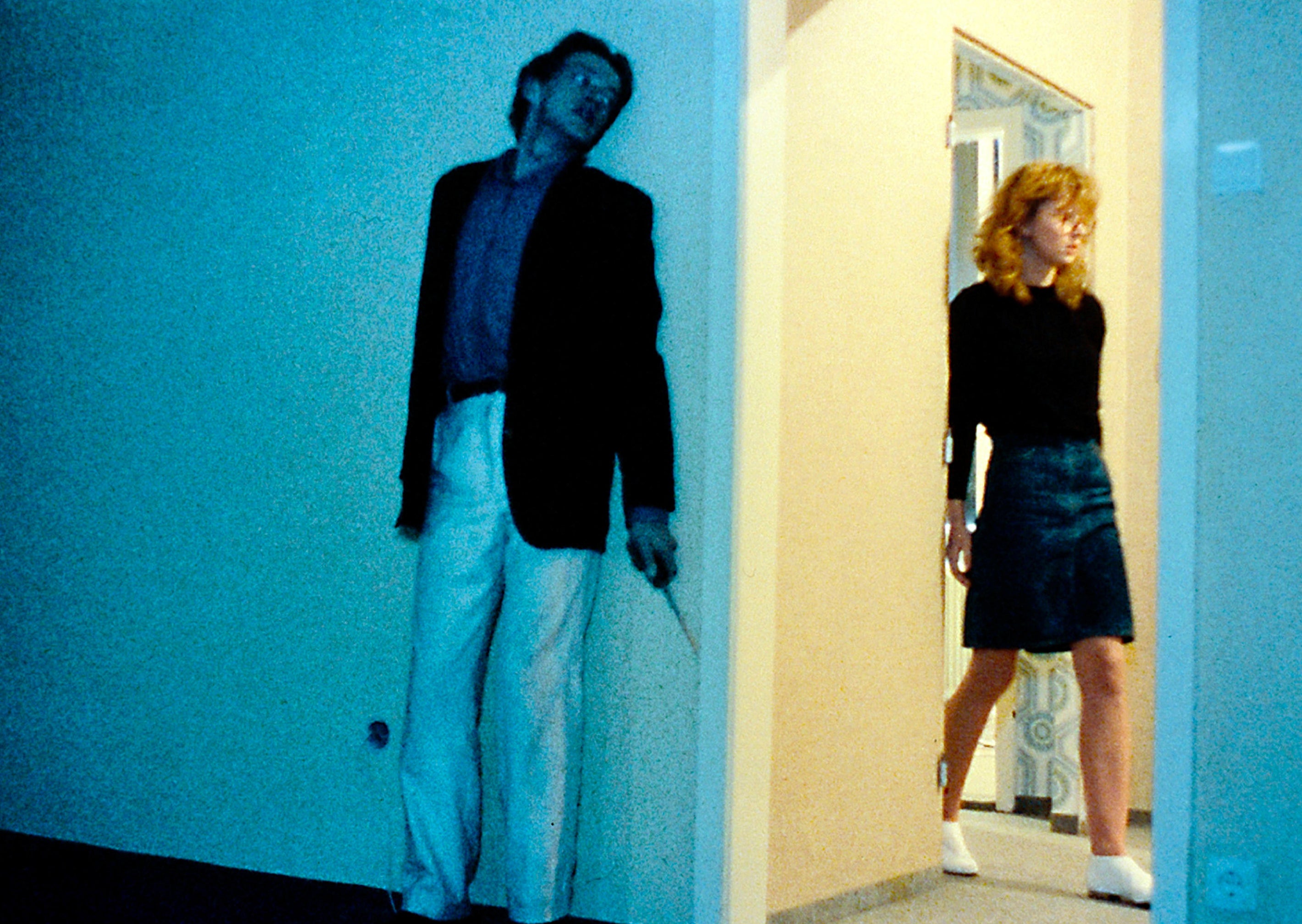 A tall, skinny and panicked man stands behind a wall while a young woman walks past a room