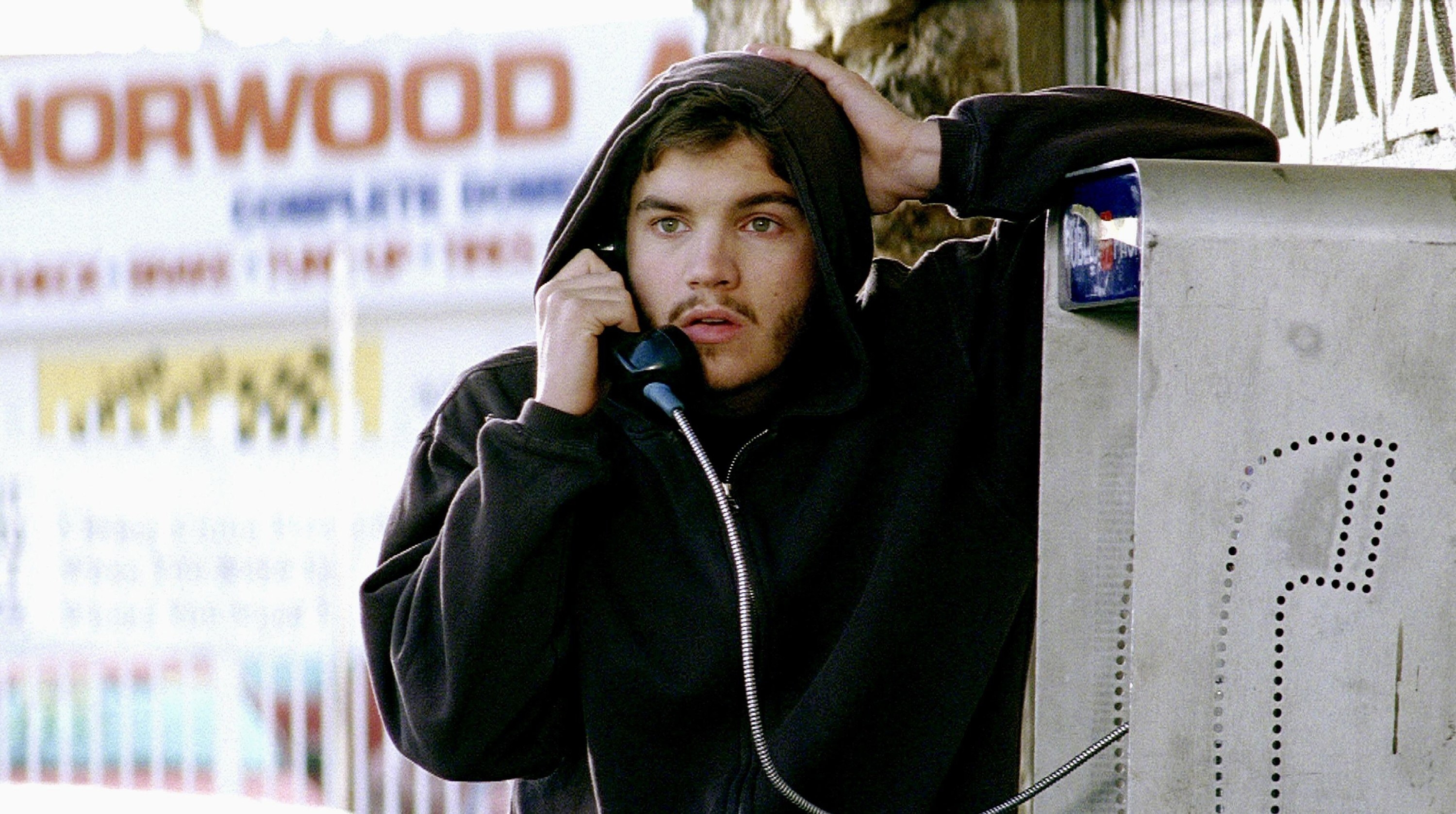 A young bearded man in a hoodie gets distressing news at a phone booth