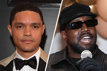 Trevor Noah wears a beige suit with a white shirt and a black tie. Kanye West wears a black leather jacket with black gloves, a black hat, and sunglasses.
