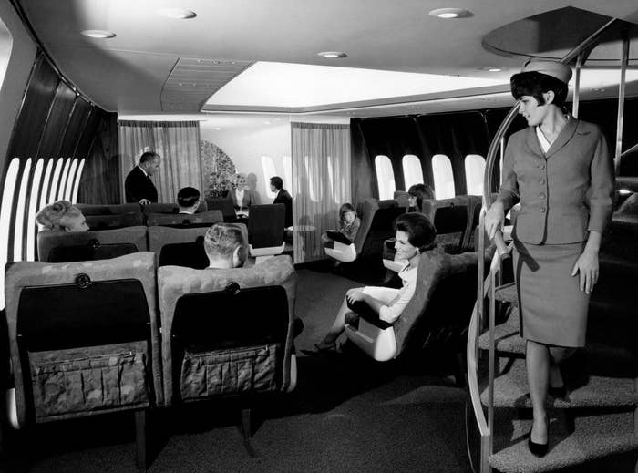 Black-and-white photo of people sitting in what looks more like a living room, with plush, far-apart seats, and a flight attendant walking down a winding staircase