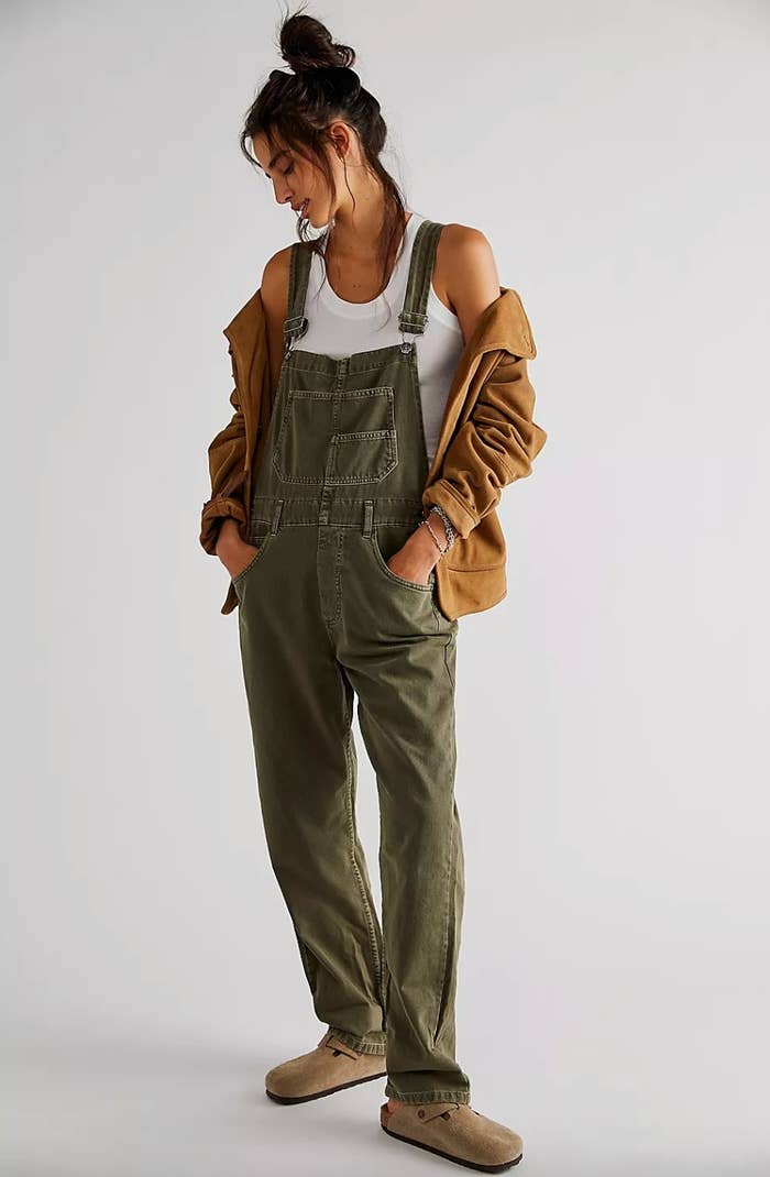 a model wearing the olive green overalls, a white tank top, a yellow zip up jacket and tan clogs