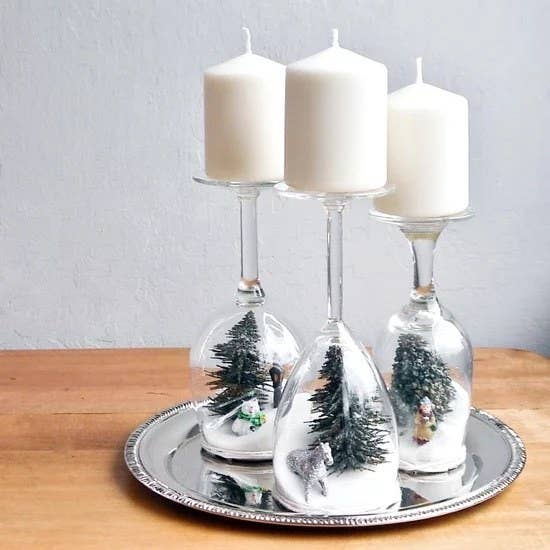 Three wine glass dioramas sit on a silver plate on a wooden counter. The wine glasses are upside down and have mini trees in the glasses, and candles sitting on the bases.