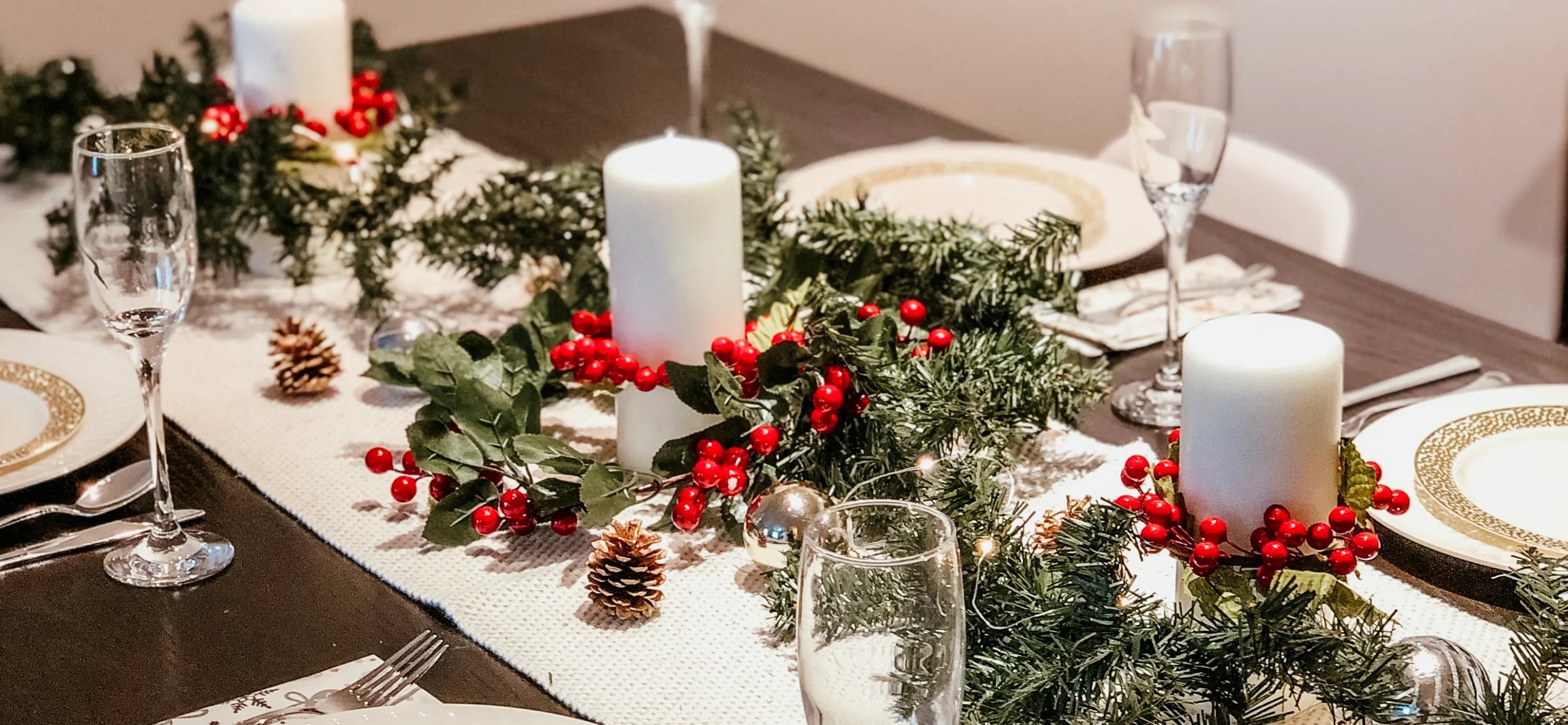 festive holiday tablescape for christmas dinner with light table runner, greenery, red berries, and pine cones with candles for a cozy feel
