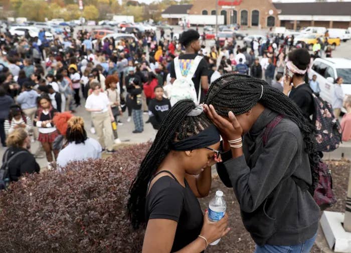Two young girls cry with their head in their hands, in the background, crowds of high schoolers stand in a parking lot outside a high school