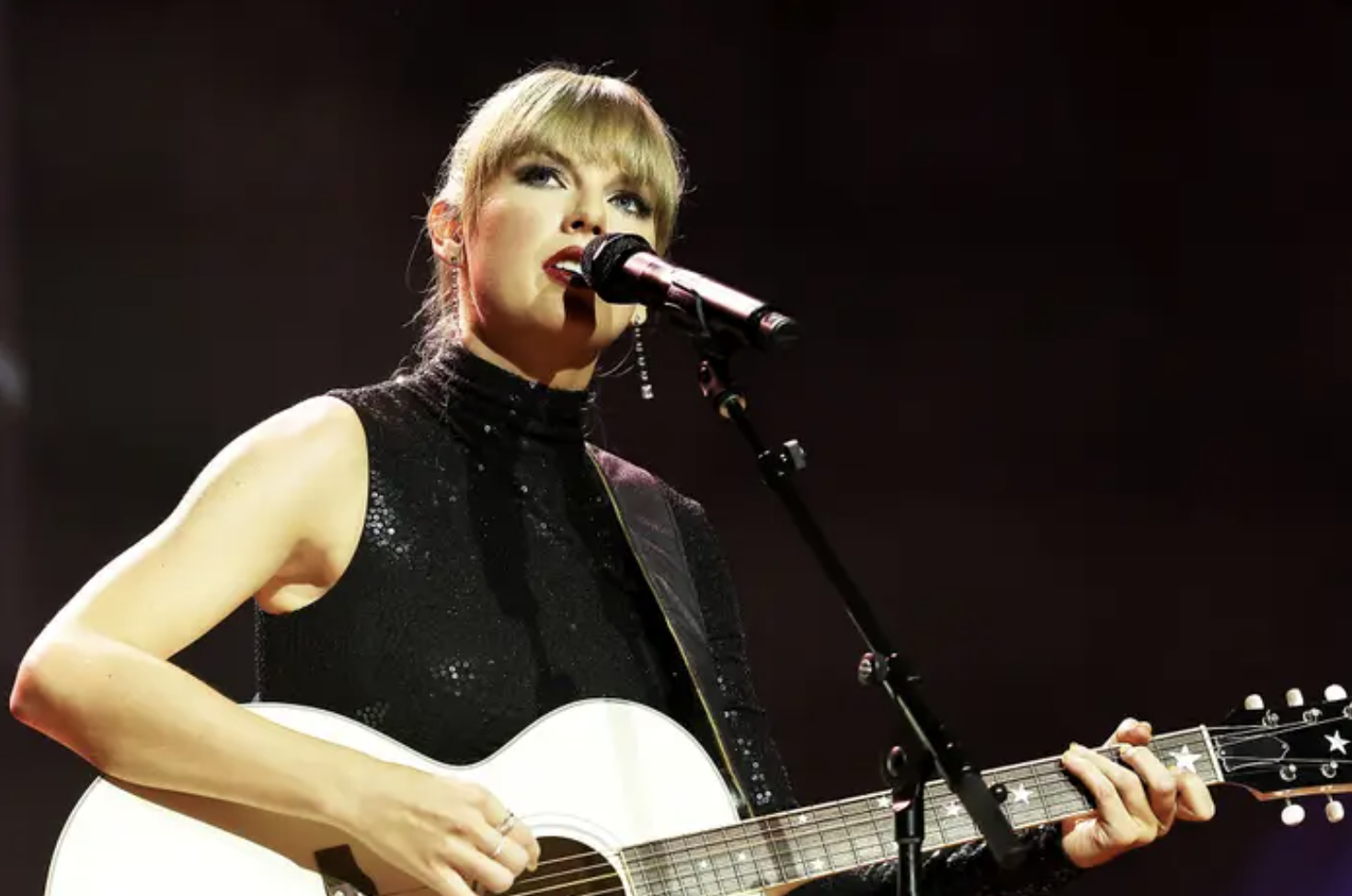 Taylor Swift stands in a sleeveless black top with a guitar in front of microphone