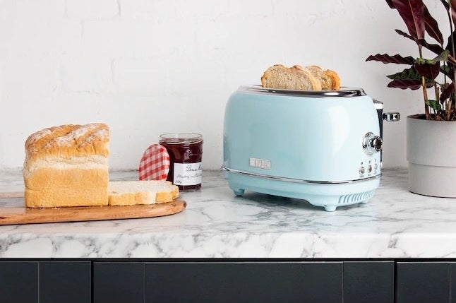 The toaster with two pieces of bread inside next to a loaf of bread and jar of jam.