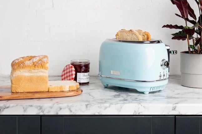 The toaster with two pieces of bread inside next to a loaf of bread and jar of jam.