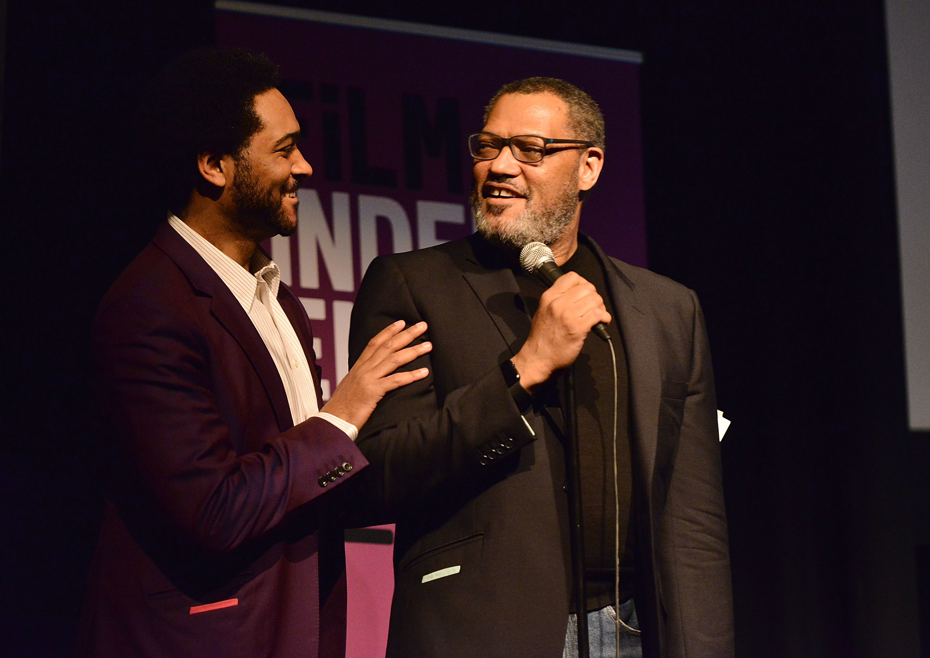 Langston onstage and smiling at his father, who&#x27;s holding a microphone and smiling at him, as he touches his shoulder