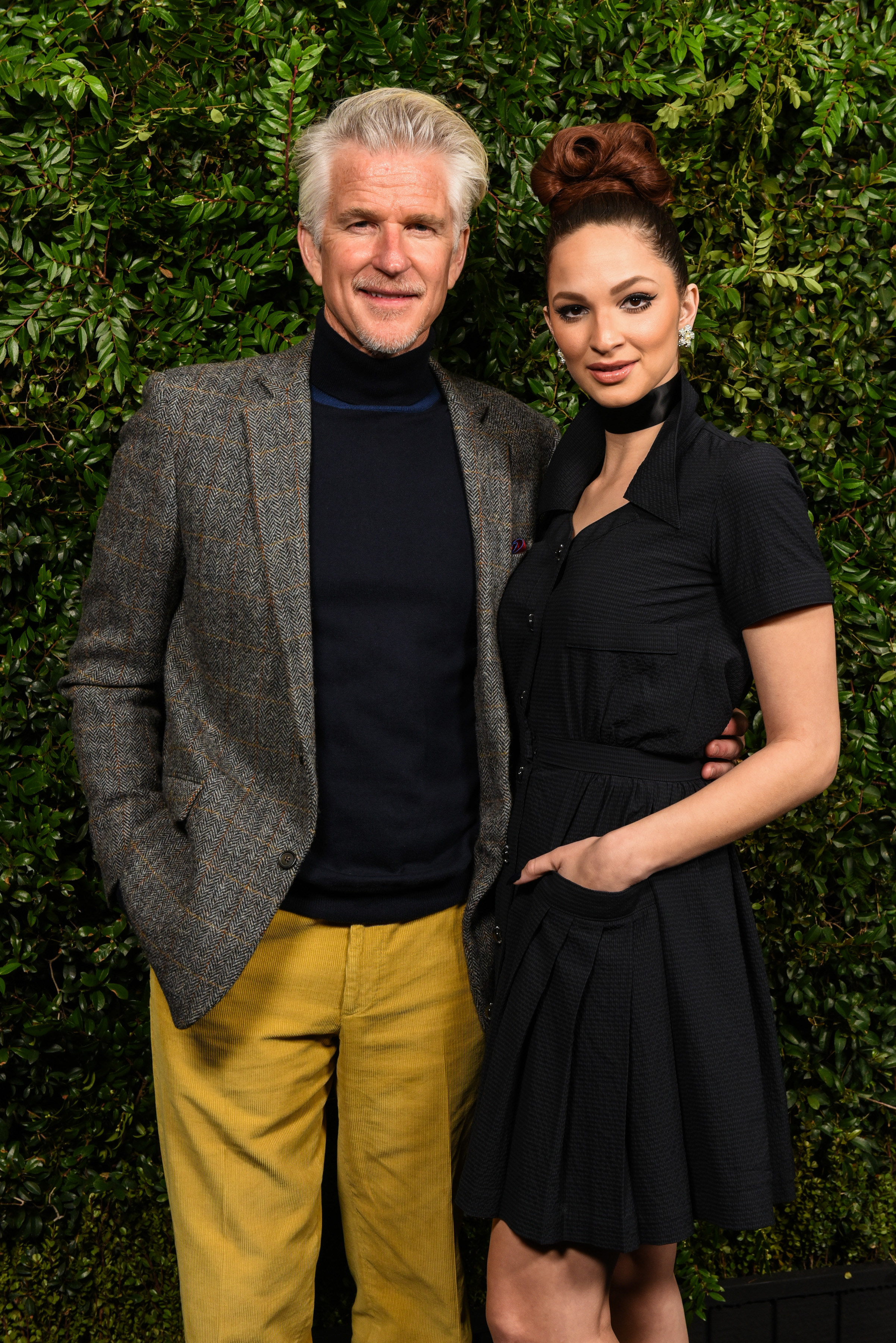 Matthew in a turtleneck with his arm around Ruby in a short dress against a backdrop of greenery