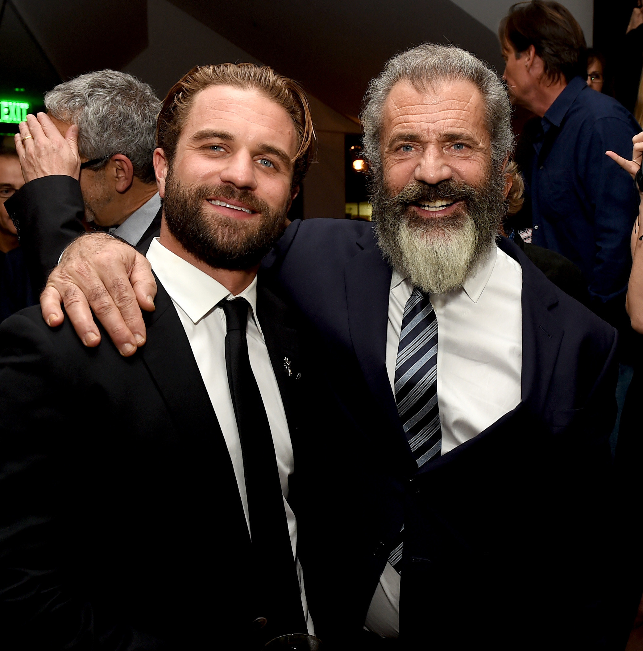 Mel smiling and with his arm around a smiling Milo&#x27;s shoulder, both wearing a suit and tie