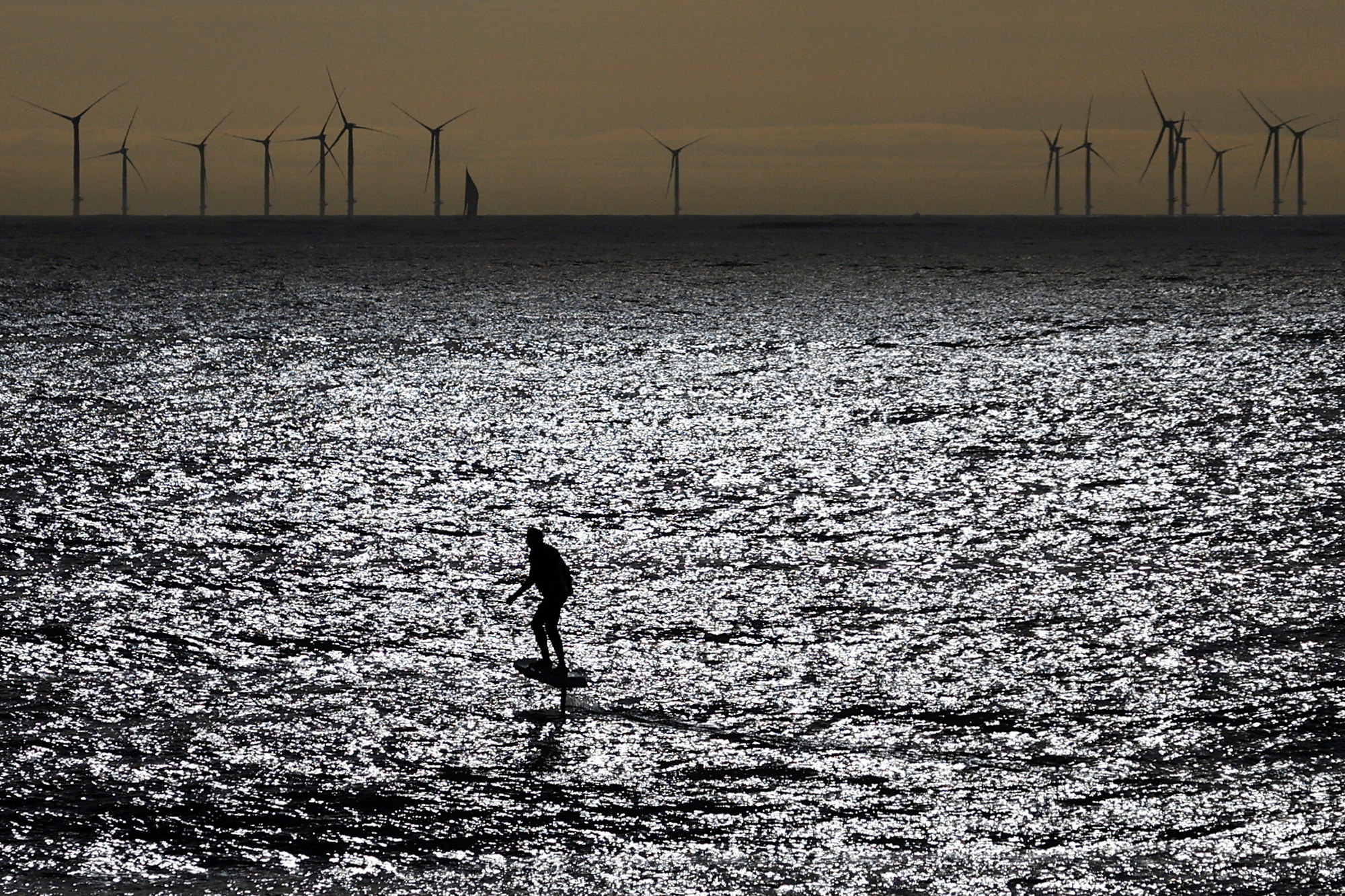 Open water at dusk; a small shadowy outline of someone on a surfboard in the middle; a line of windmills in the background