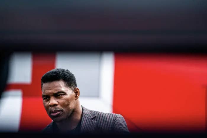 Herschel Walker in a gingham suit against a red background; the top part of the shot is covered by a dark wall that&#x27;s out of focus