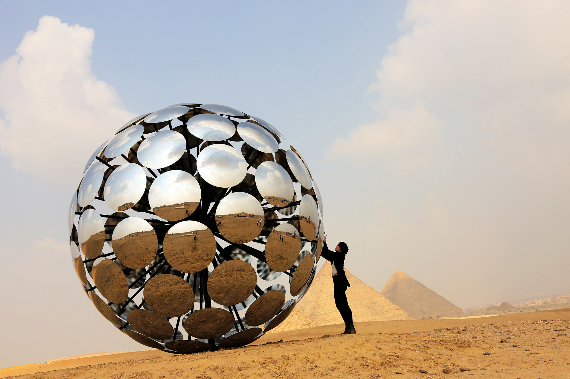 Someone holds up a giant silver reflective ball, in the background are the Giza pyramids