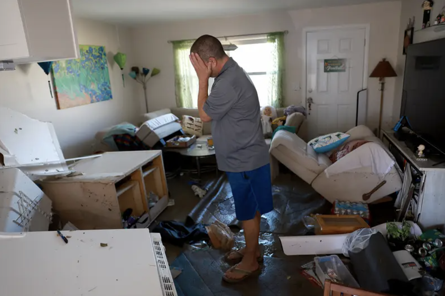 a man covers his face in emotion and stands in a room of his destroyed belongings after a storm