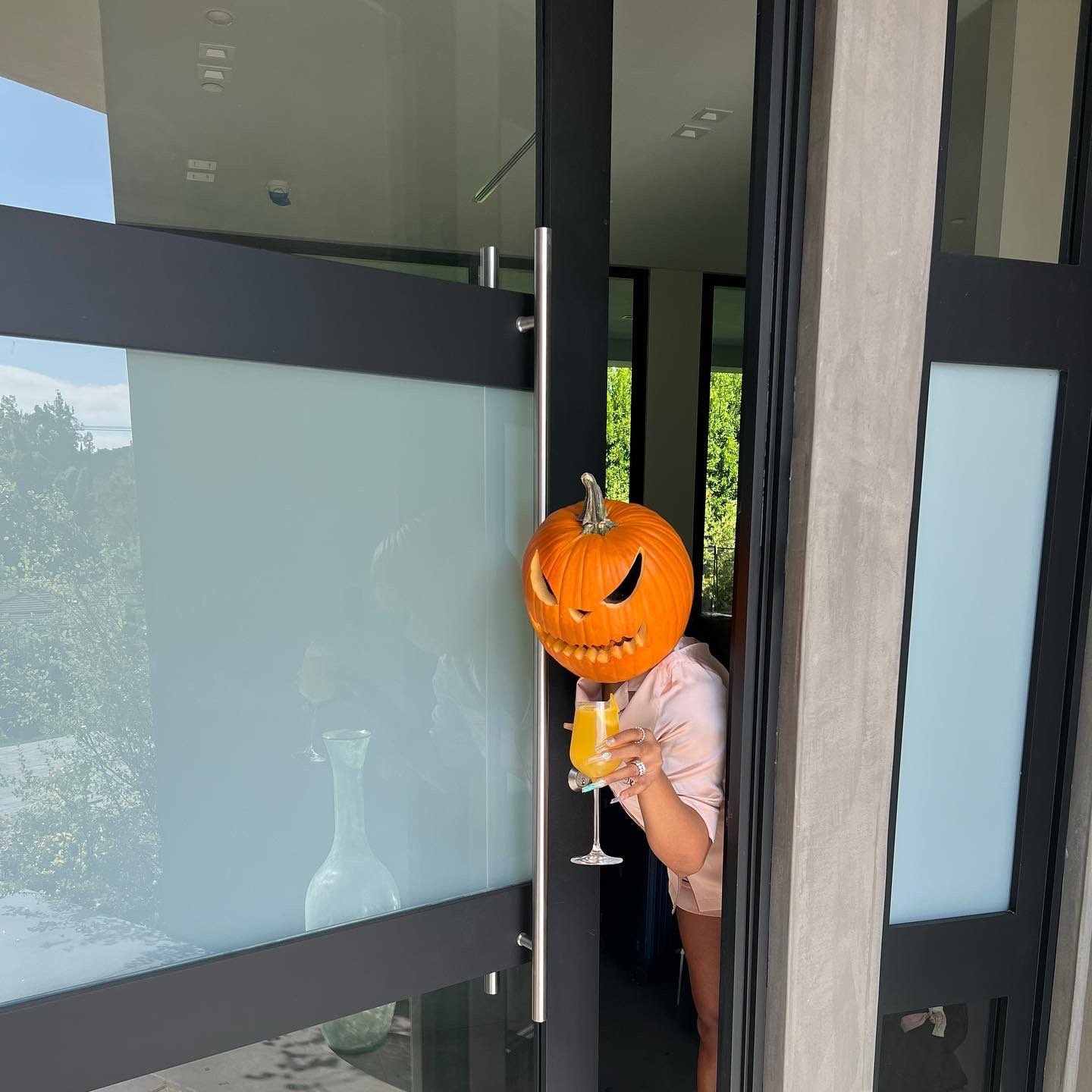 Megan wears a pumpkin on her head and opens her front door holding a glass