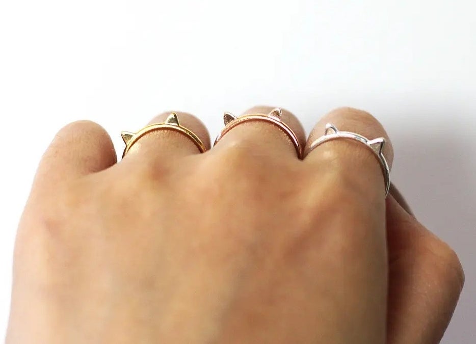 a hand with three delicate cat rings on separate fingers, in gold, rose gold, and silver, respectively
