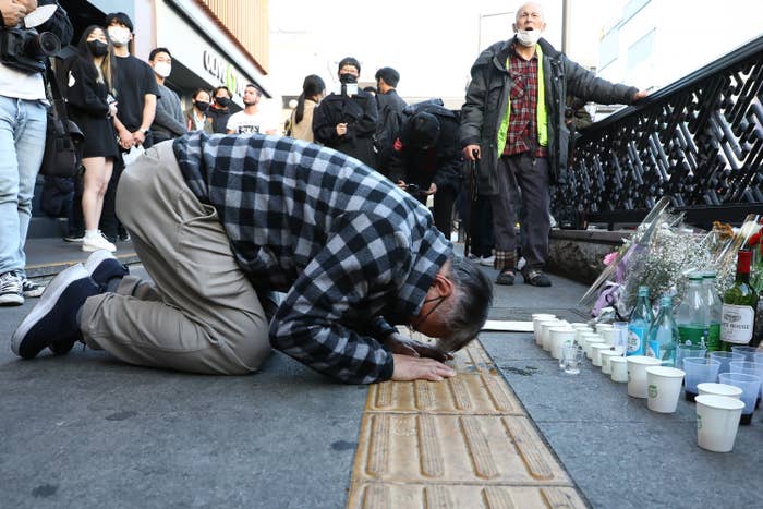A man kneels on the ground in front of a street memorial as people look on