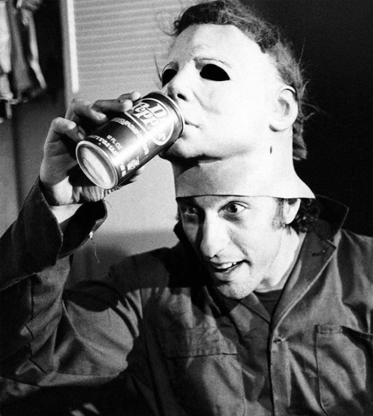 black and white photo of nick castle with a mask on top of his head, he&#x27;s having the mask pretend to drink dr pepper