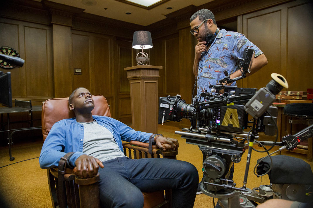 daniel kaluuya sits leaning back in a chair in front of a film camera, jordan peele stands over him looking pensive