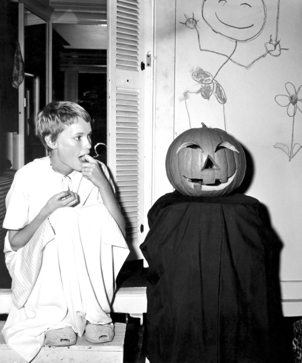 mia farrow on the left crouching with her hand in her mouth; on the right is a jack o lantern sitting on top of a black sheet