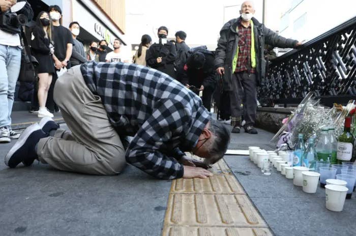 a man in a checkered shirt bows with his forehead touching the ground at a memorial outside