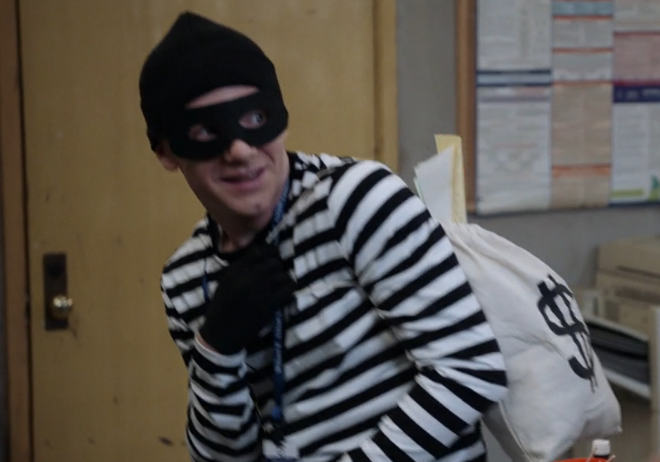 Jacob is wearing a black-and-white striped long-sleeved shirt, a beanie, gloves, and an eye mask as he carries a bag with the U.S. dollar sign