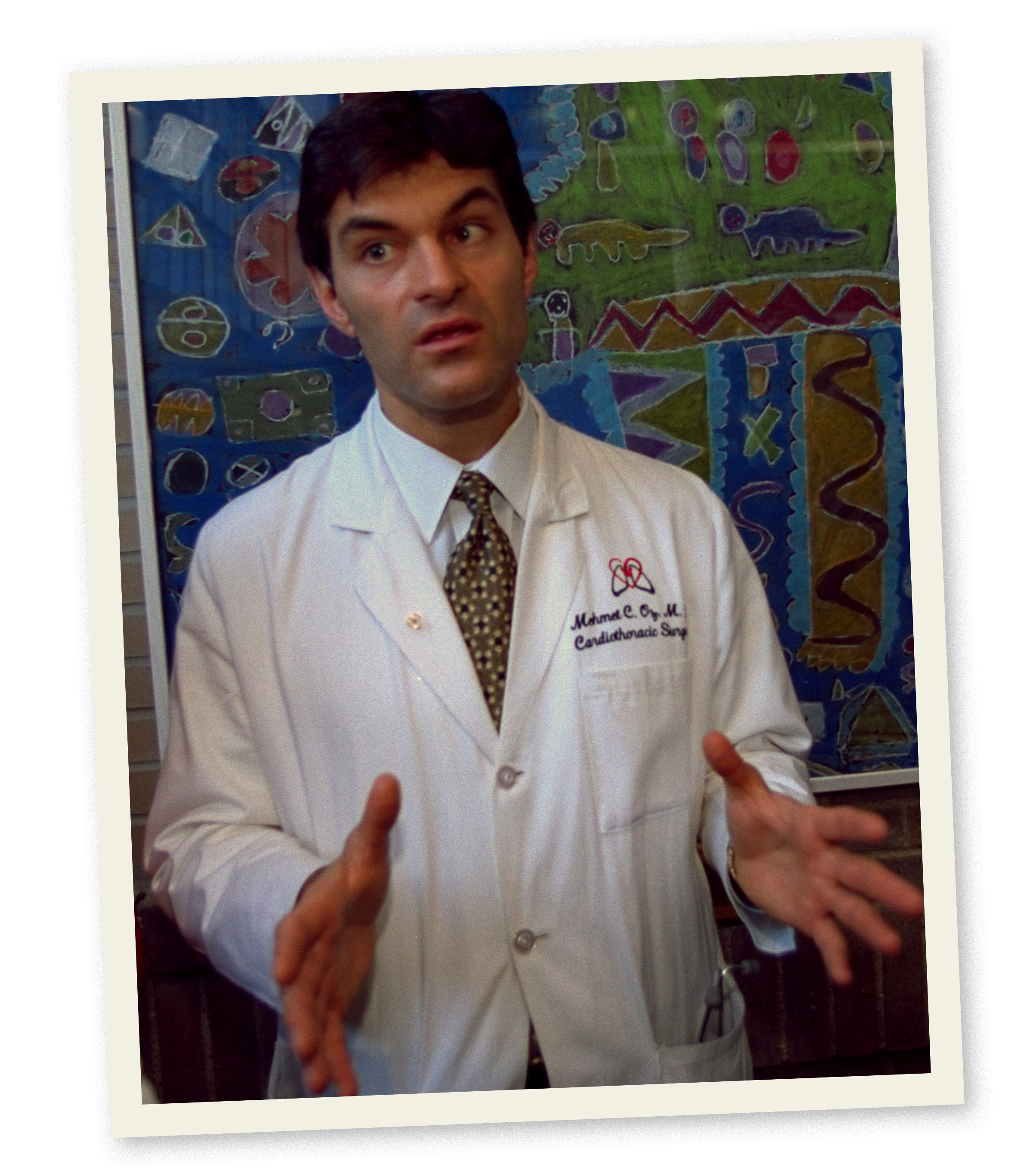Dr Oz gestures with his hands in front of him while wearing a white doctor&#x27;s jacket and standing in front of a wall with simple children&#x27;s illustrations