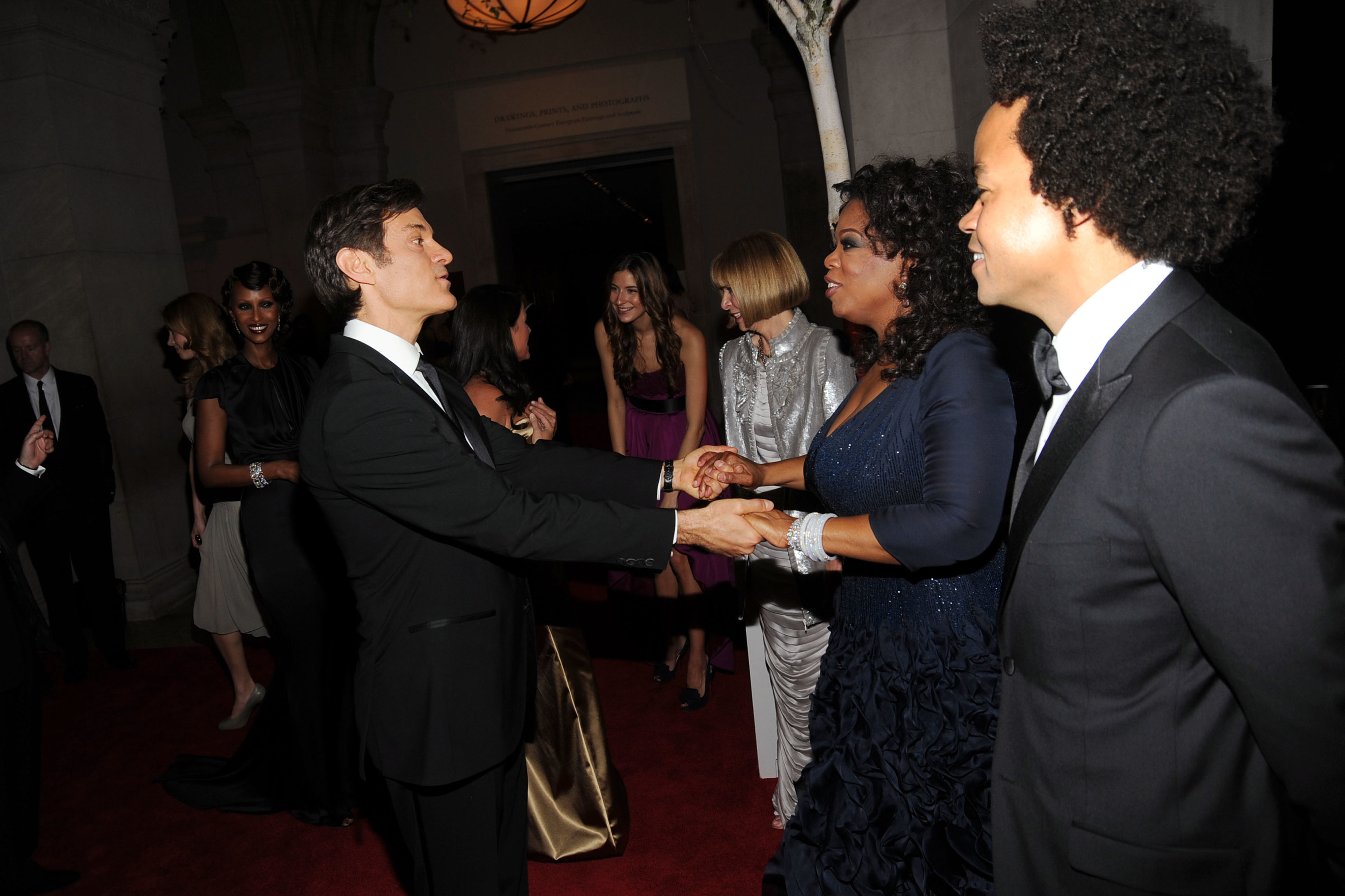 Oz and Oprah at a black-tie event, holding each others&#x27; hands at a distance and surrounded by others dressed formally at an indoor venue