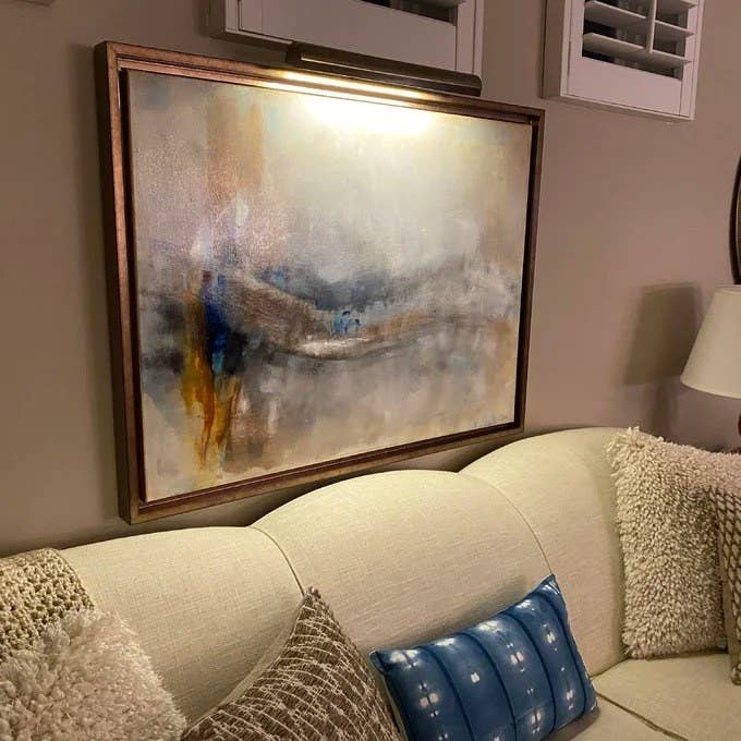 the overhead light illuminating an abstract painting hung over a beige couch