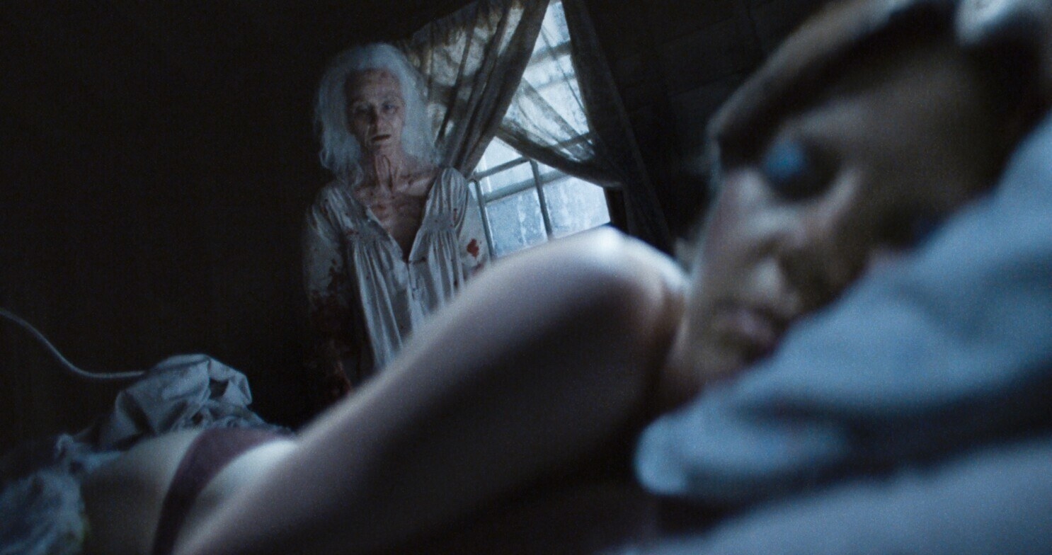 Maxine Minx (Mia Goth) laying in bed asleep in a tight shot. Behind is Pearl (also Mia Goth), an elderly woman, staring at Minx asleep.