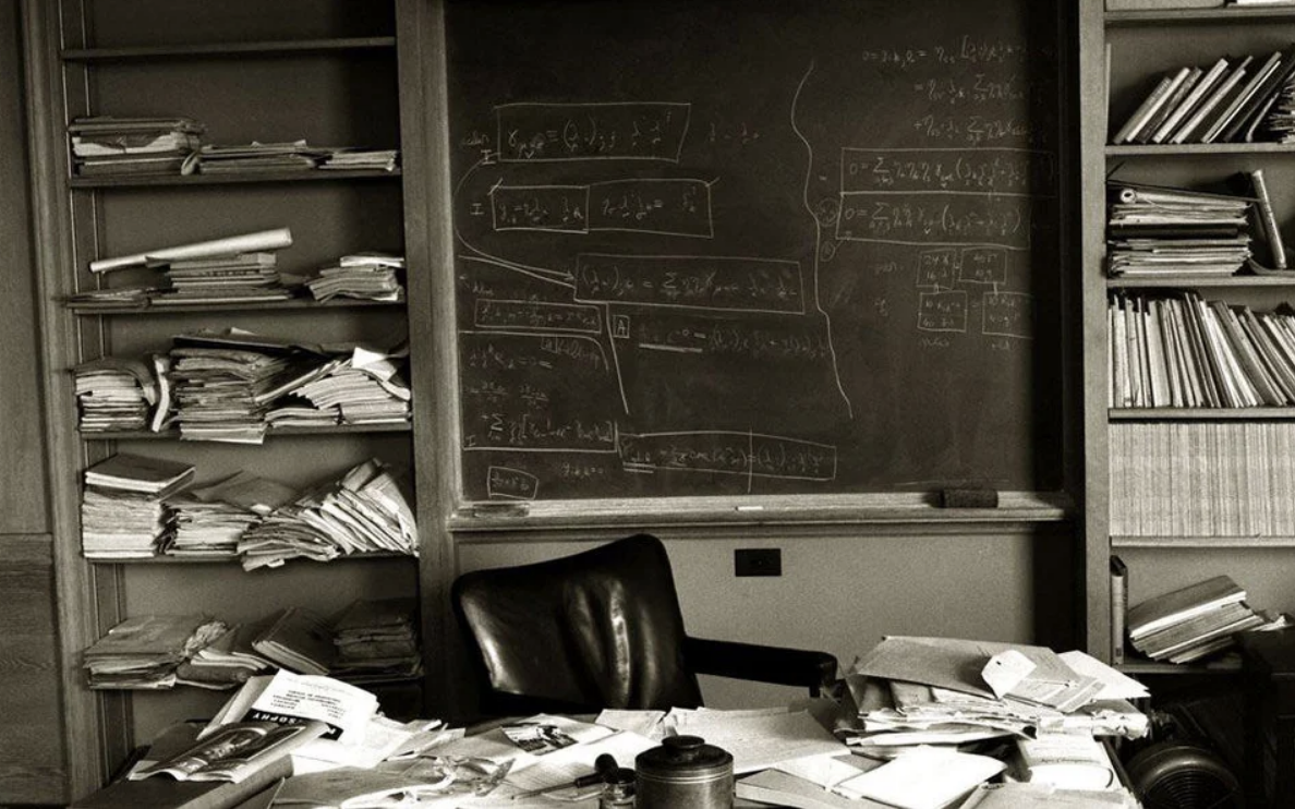 A desk with many papers on it in front of a chair and a blackboard with writing on it, and magazines, journals, and papers on shelves on either side