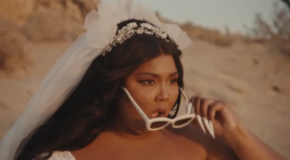 Lizzo in a wedding dress taking her sunglasses off and opening her mouth in surprise