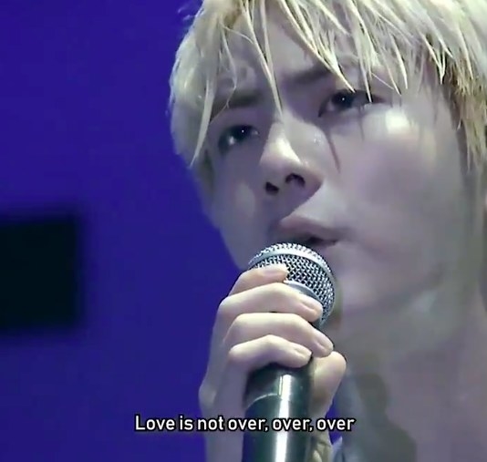 Close-up of Jin with bleached blond hair singing into a microphone with the caption &quot;Love is not over, over, over&quot;
