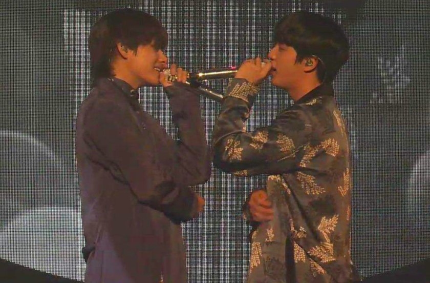 V and Jin sing into microphones while facing and looking at each other and smiling, standing close together