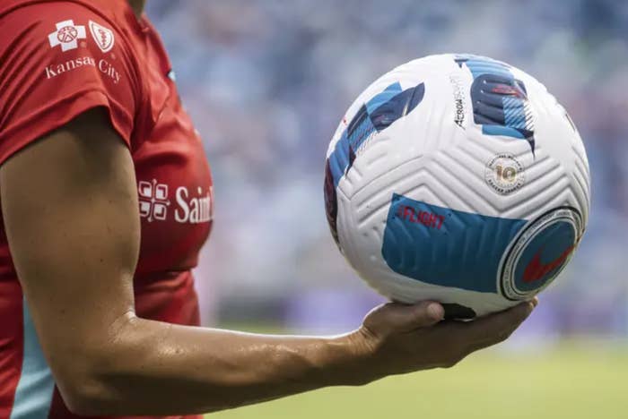 a woman wearing a jersey and holding a soccer ball on the field; the image is zoomed in on the ball and her torso
