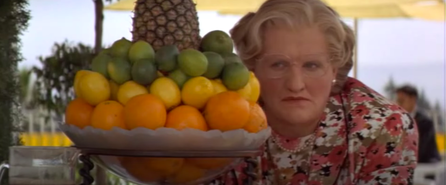 A man dressed as an elderly woman hides behind a bowl of fruit