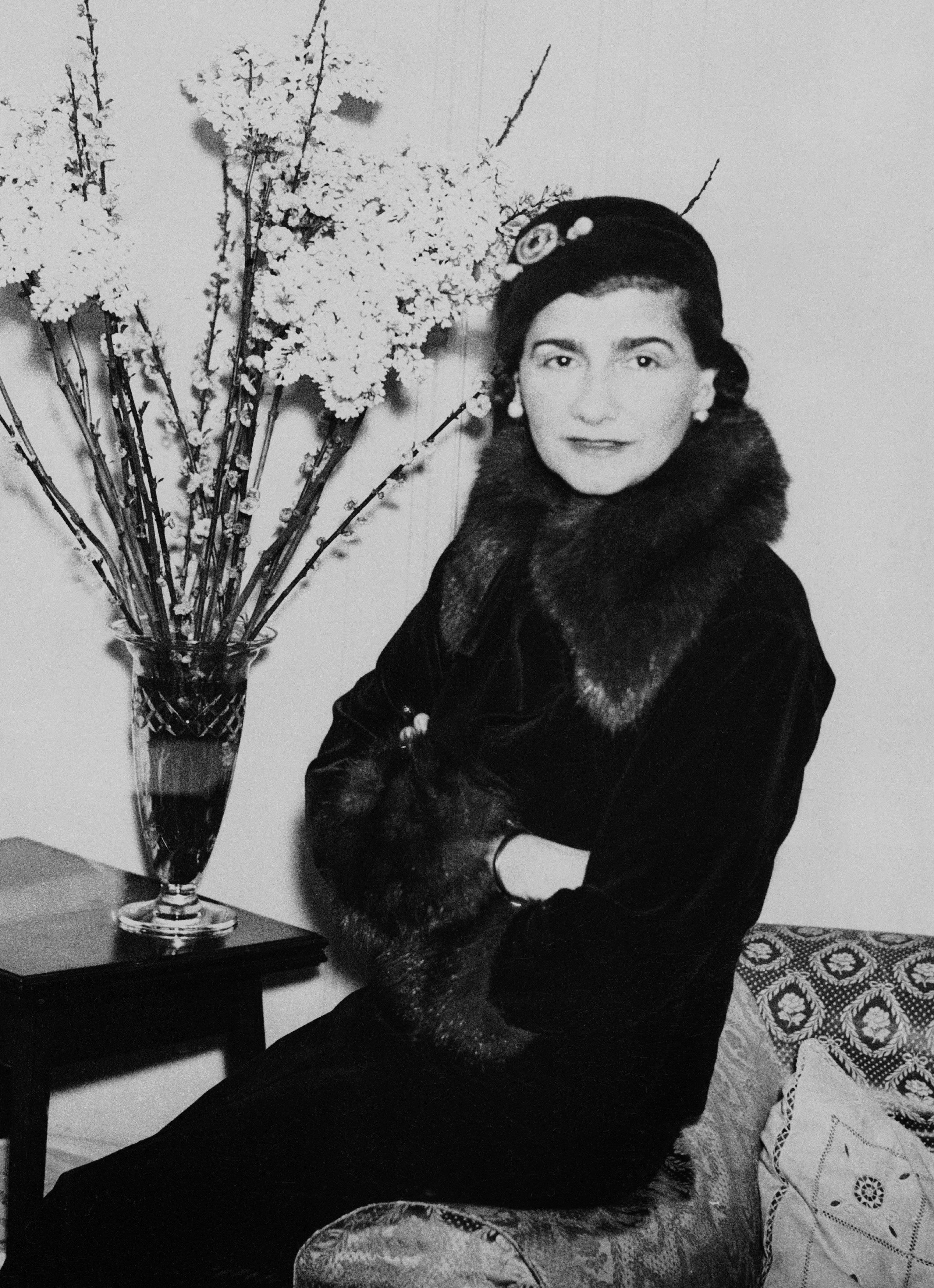 Black-and-white photo of Coco perching on the arm of a couch and wearing a fur coat with her arms folded