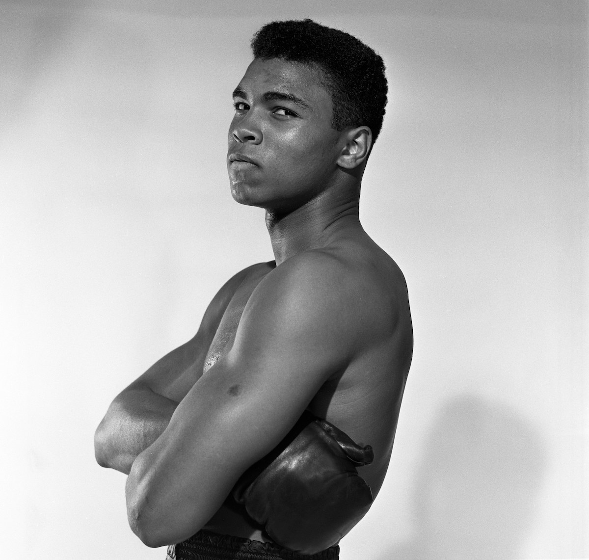 A black-and-white photo of a bare-chested Ali with his arms folded and wearing boxing gloves
