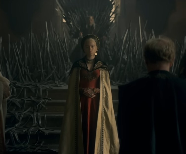Milly Alcock as a young Rhaenyra Targaryen standing in front of the iron throne and the King as people swear fealty to her
