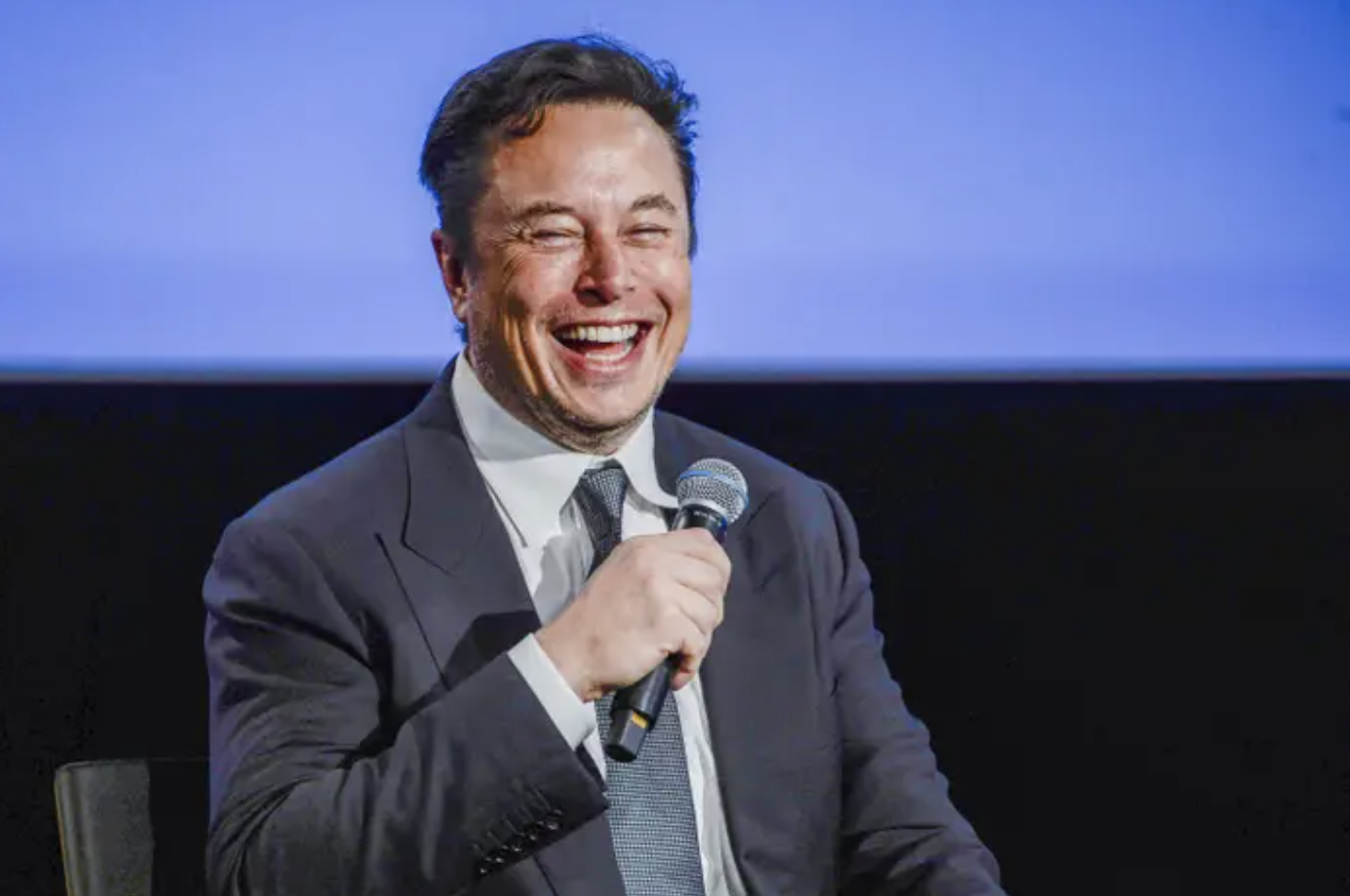 Elon Musk wearing a suit, holding a microphone and laughing