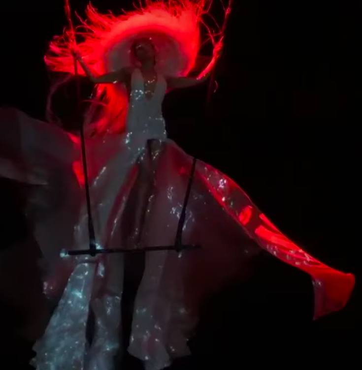 Model wearing a gauzy costume on a trapeze against a dark background