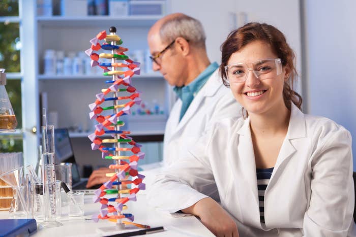A scientist smiles at the camera as she sits next to a model of DNA as another scientist works behind her