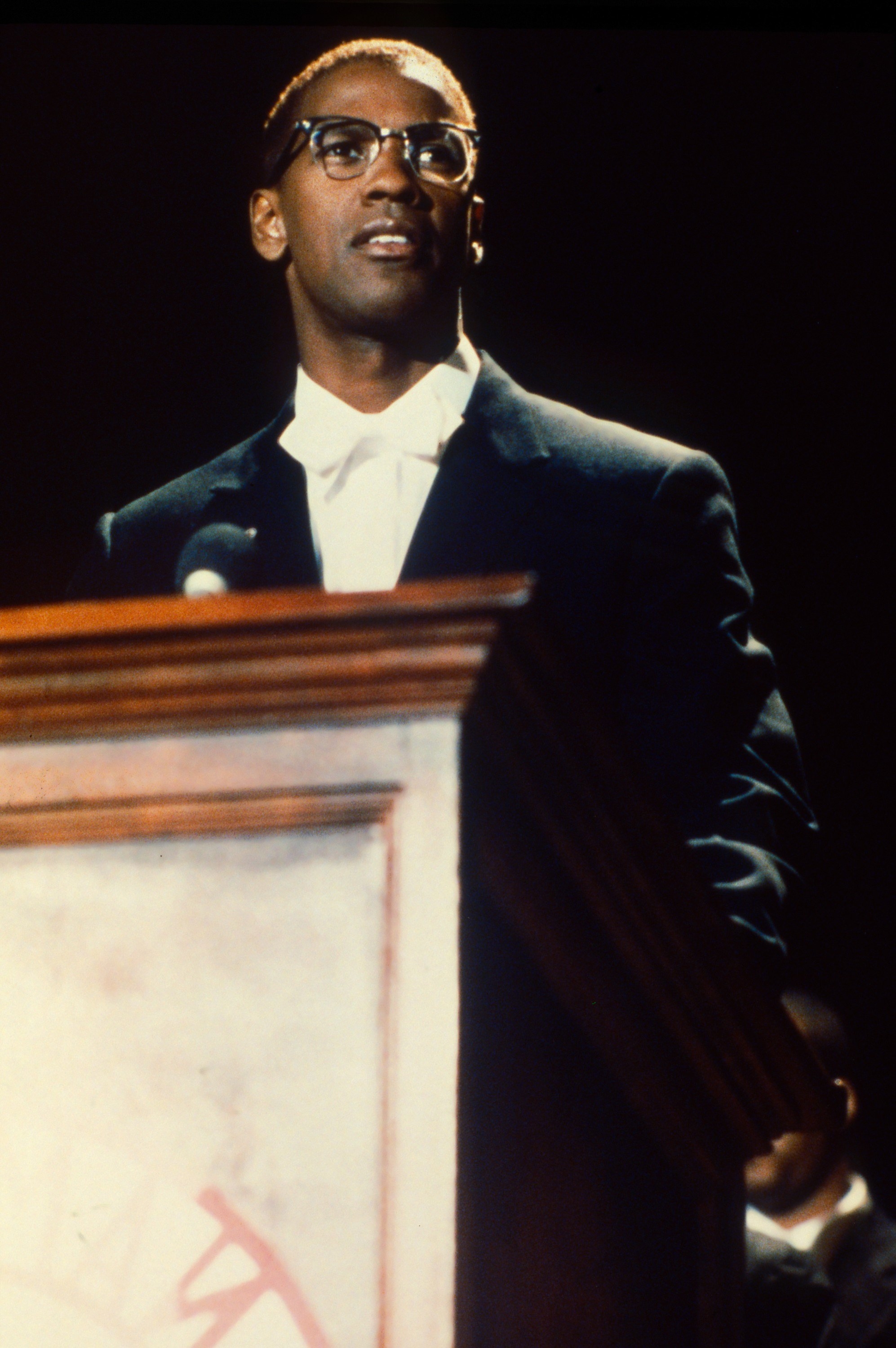 Denzel as Malcolm wearing glasses and a suit and bow tie and standing at a podium