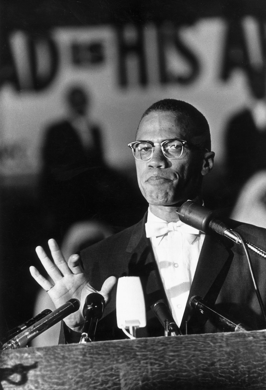 Black-and-white photo of Malcolm wearing glasses and a suit and bow tie and standing at a podium