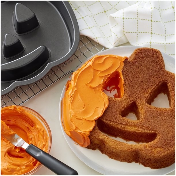 Jack-o-lantern cake pan next to cake being iced with orange frosting