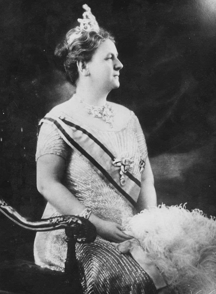 A black-and-white photo of a woman sitting in a high-back chair and wearing a crown and a short-sleeved dress