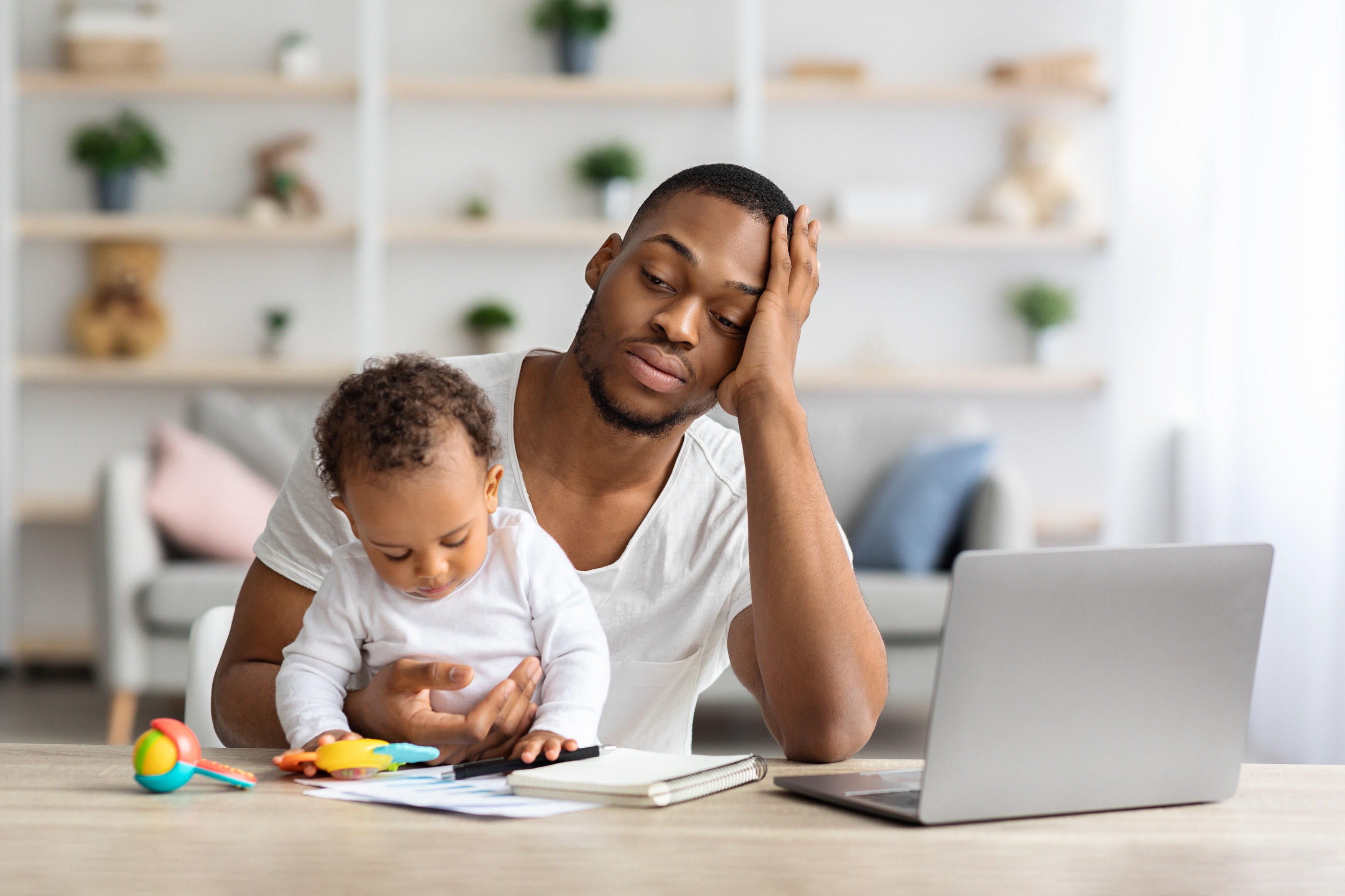 A father looks stressed as he watches his baby and works on his laptop