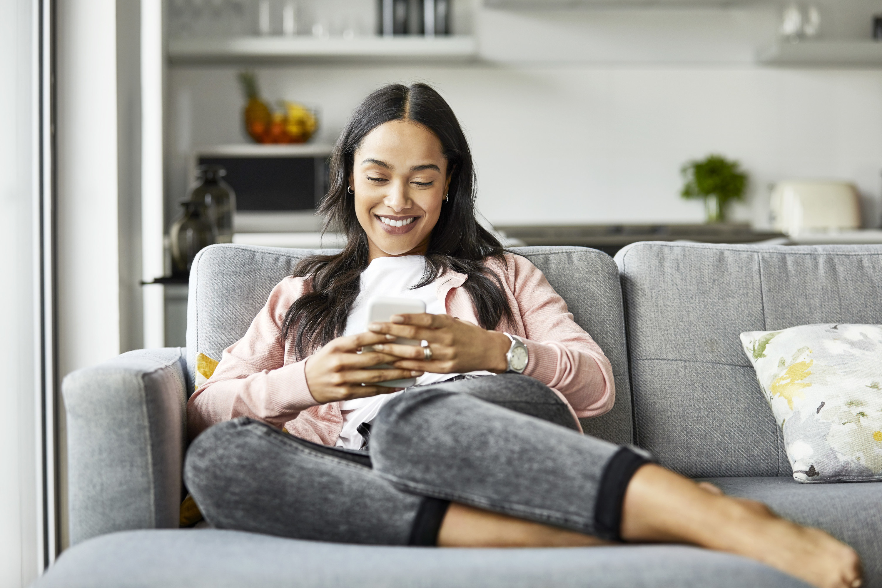 A woman smiles as she sits on her couch and checks her phone