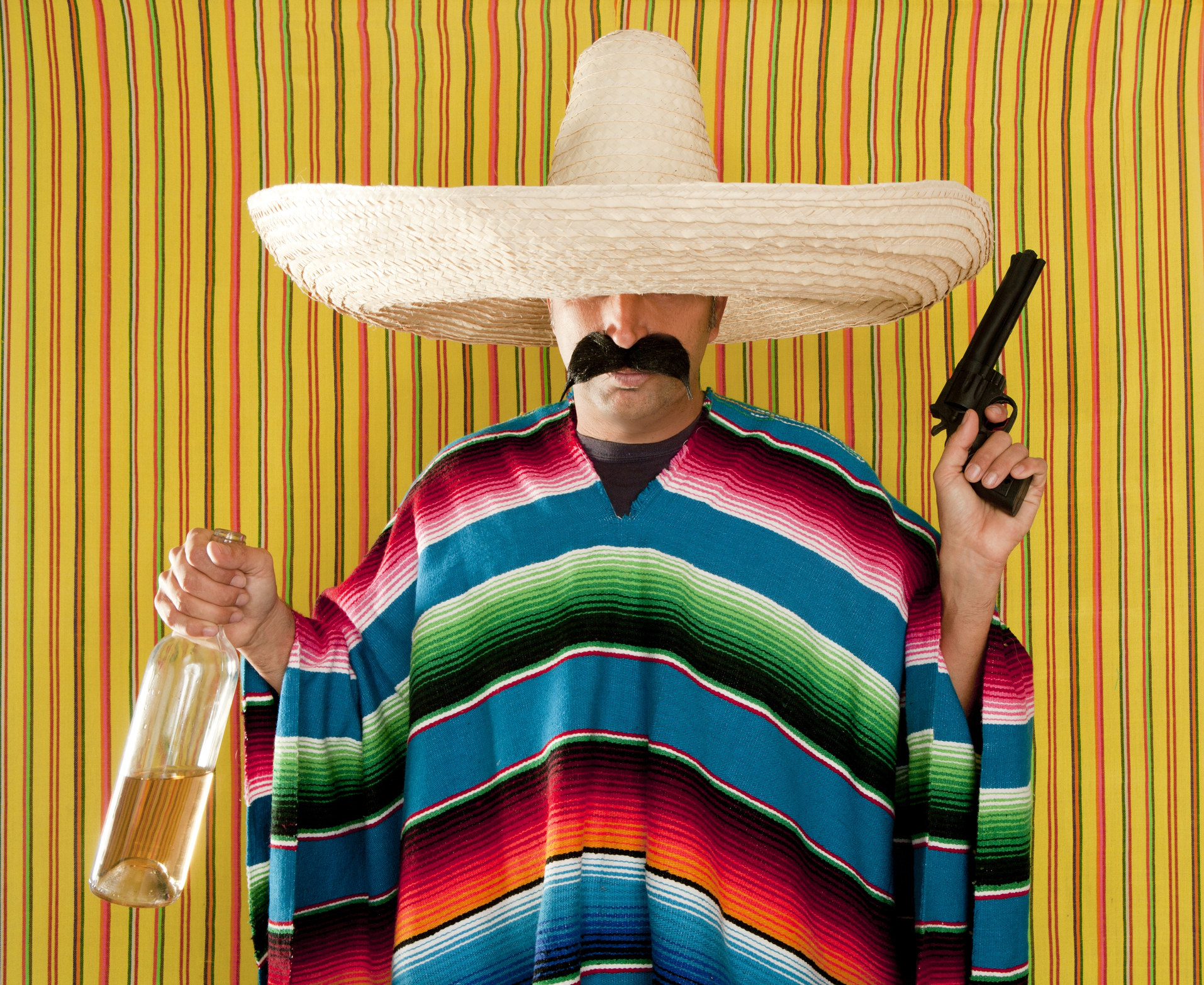 A man wearing a fake mustache, a poncho, a sombrero, and holding a gun and tequila